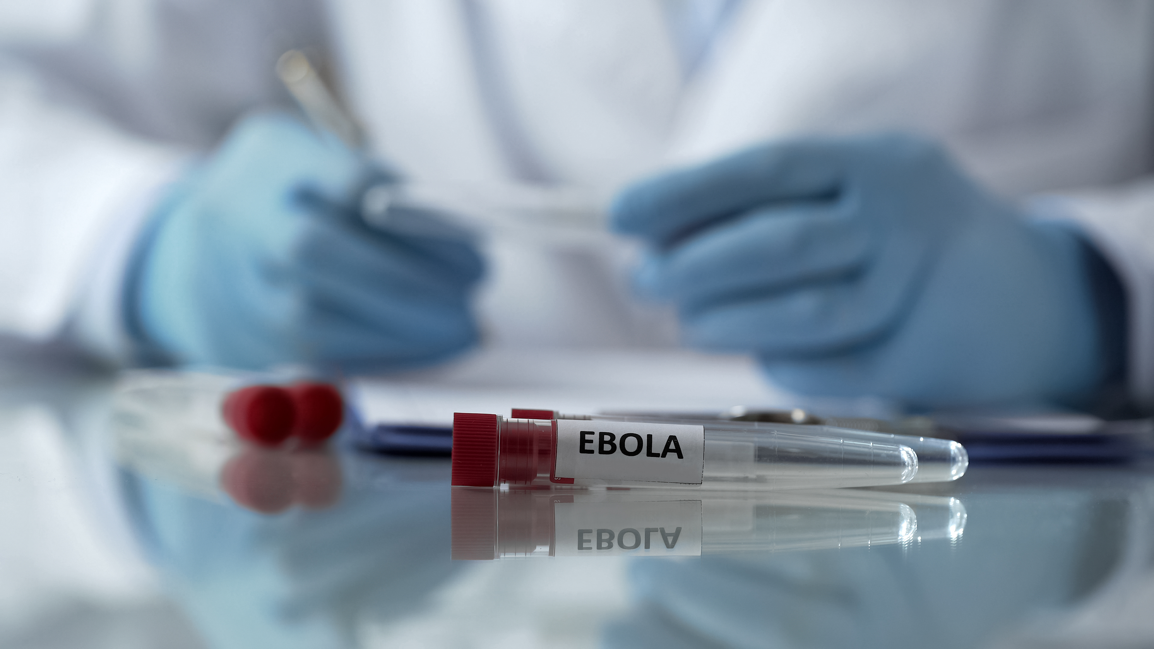 Imagem de um ambiente laboratorial com foco em um tubo de ensaio identificado com a palavra “EBOLA”. O tubo está apoiado sobre uma superfície reflexiva, enquanto ao fundo aparece uma profissional da saúde desfocada, usando luvas azuis e manipulando materiais médicos. A composição transmite clima de alerta epidemiológico e investigação científica.