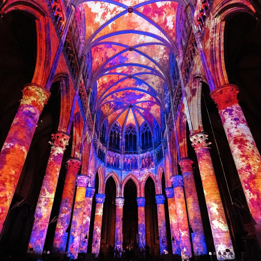Interior de catedral iluminado com projeções coloridas em tons vibrantes, destacando a arquitetura gótica.