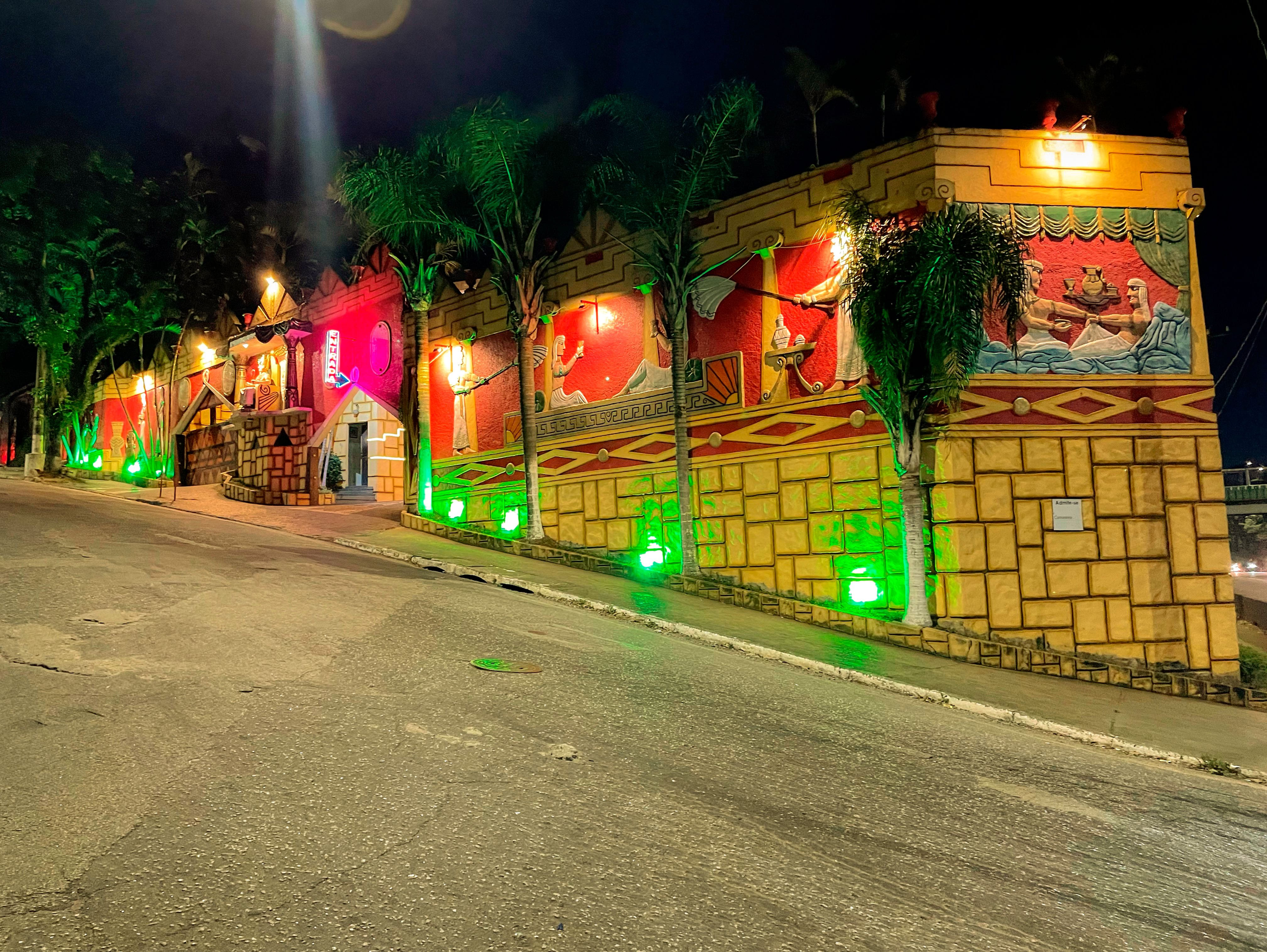 Fachada de motel com tem&aacute;tica eg&iacute;pcia &agrave; noite, iluminada por luzes verdes e amarelas. A constru&ccedil;&atilde;o tem paredes amarelas com blocos texturizados e murais vermelhos retratando figuras eg&iacute;pcias. Palmeiras e arbustos verdes complementam a decora&ccedil;&atilde;o, com uma rua asfaltada em primeiro plano