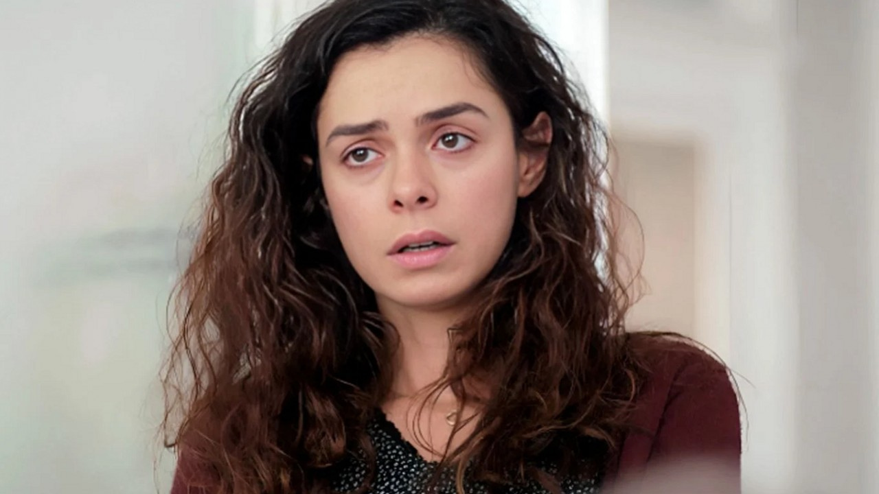 Close no rosto de uma jovem de pele clara e cabelos longos, escuros e cacheados, levemente desalinhados. Ela est&aacute; em um ambiente interno claro, com fundo desfocado em tons brancos e bege. A mulher olha para frente com express&atilde;o triste e preocupada. Seus olhos parecem marejados, como se estivesse prestes a chorar, e sua boca est&aacute; ligeiramente aberta, transmitindo surpresa, medo ou ang&uacute;stia. Ela veste um casaco escuro em tom vinho sobre uma blusa preta com pequenos detalhes claros. A ilumina&ccedil;&atilde;o suave destaca seu rosto de maneira natural, refor&ccedil;ando o clima emocional e dram&aacute;tico da cena.