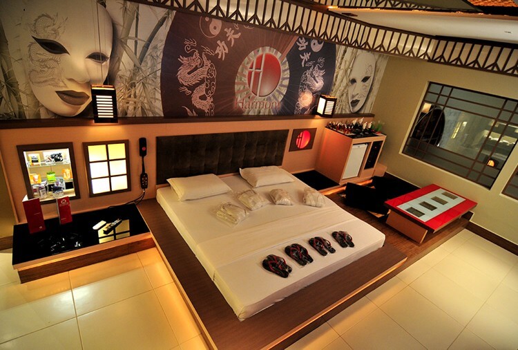 Quarto de motel com decora&ccedil;&atilde;o asi&aacute;tica, cama de casal branca com chinelos pretos e roup&otilde;es, painel com m&aacute;scaras e drag&otilde;es, e mesa de centro vermelha