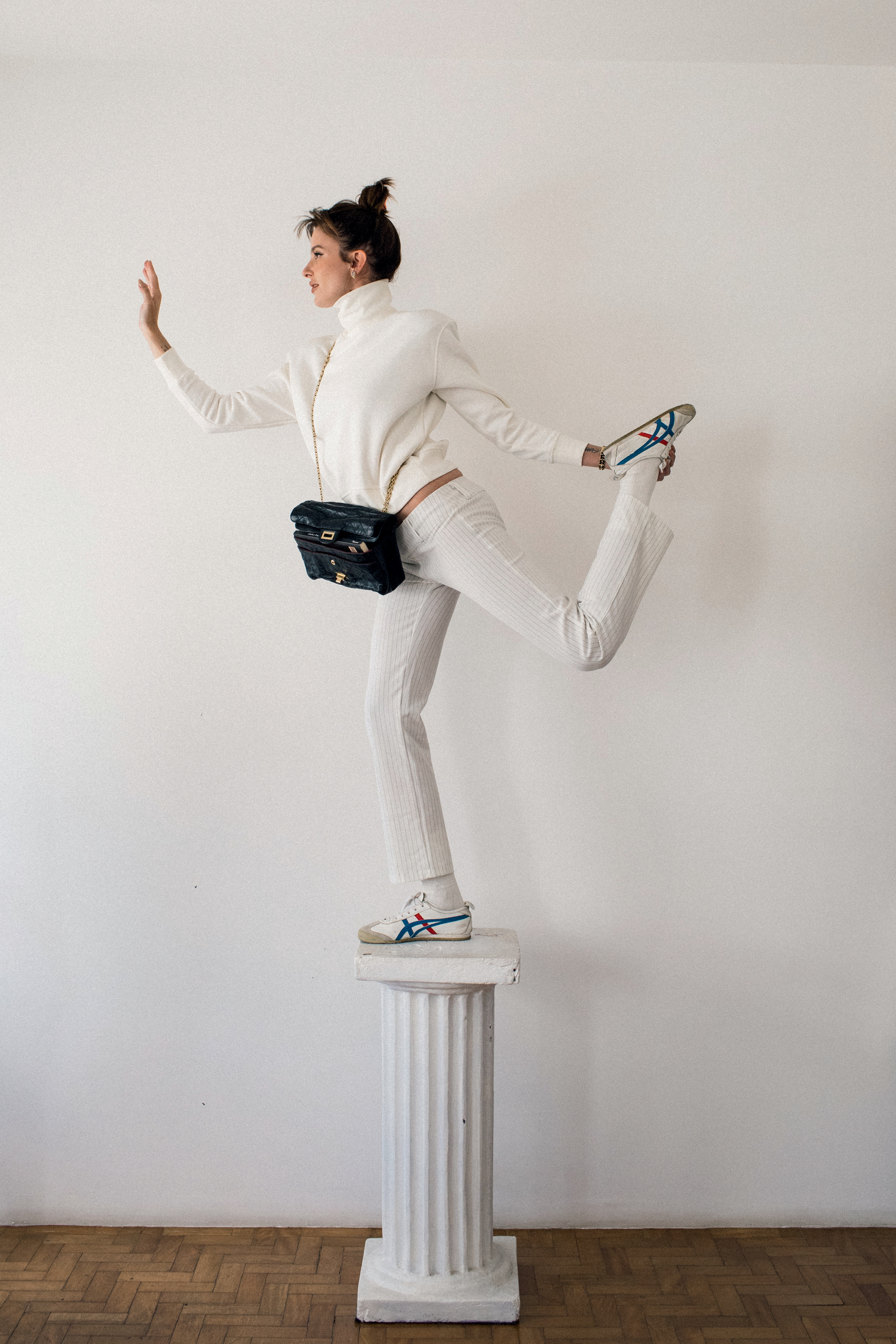 Foto de Verena Figueiredo. Na imagem, ela aparece com um moletom branco, uma cal&ccedil;a branca, uma bolsa preta de couro da Chanel e t&ecirc;nis branco. Ela est&aacute; em cima de uma pilastra, em p&eacute;, segurando um dos p&eacute;s com a m&atilde;o e com a outra para a frente