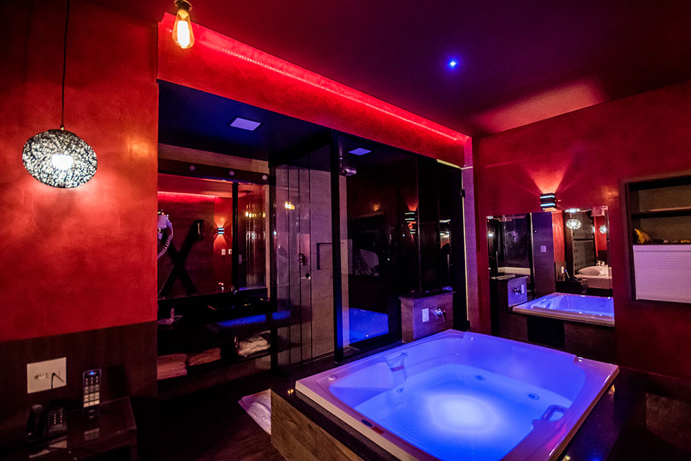 Quarto de motel com paredes vermelhas, ilumina&ccedil;&atilde;o ambiente em tons de vermelho e azul, e duas banheiras de hidromassagem com luzes azuis. Ao fundo, espelhos refletem um ambiente com uma cruz decorativa na parede e uma cama. Um telefone e controle remoto est&atilde;o em uma mesa lateral.