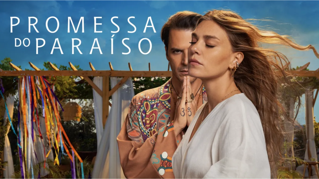 Imagem promocional com clima leve e espiritual ao ar livre. Em primeiro plano, &agrave; direita, aparece uma mulher jovem de pele clara e cabelos longos castanho-claros esvoa&ccedil;ando ao vento. Ela est&aacute; de olhos fechados, com express&atilde;o serena e introspectiva, como se estivesse meditando ou sentindo a brisa. Usa roupa branca de tecido leve, brincos pequenos e alguns an&eacute;is. Atr&aacute;s dela, parcialmente escondido, est&aacute; um homem jovem de pele clara e cabelos castanhos penteados para tr&aacute;s. Ele olha diretamente para a frente com express&atilde;o intensa e misteriosa. Usa uma camisa estampada bem colorida, com desenhos psicod&eacute;licos em tons vibrantes. As m&atilde;os dele est&atilde;o unidas em posi&ccedil;&atilde;o de ora&ccedil;&atilde;o, pr&oacute;ximas ao rosto, e em um dos dedos h&aacute; o s&iacute;mbolo &ldquo;Om&rdquo; tatuado. Ao fundo, h&aacute; uma estrutura de madeira decorada com tecidos brancos e v&aacute;rias fitas coloridas penduradas, lembrando um espa&ccedil;o de retiro espiritual ou cerim&ocirc;nia ao ar livre. O c&eacute;u azul claro refor&ccedil;a a sensa&ccedil;&atilde;o de tranquilidade. No canto superior esquerdo da imagem est&aacute; escrito, em letras brancas grandes: &ldquo;PROMESSA DO PARA&Iacute;SO&rdquo;.