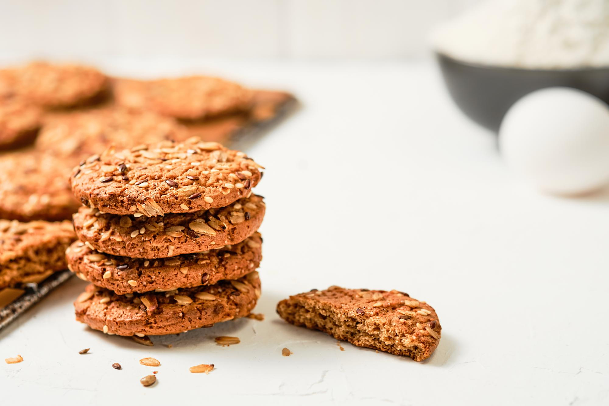 A imagem apresenta biscoitos crocantes empilhados, preparados com sementes variadas como linha&ccedil;a, aveia e gergelim