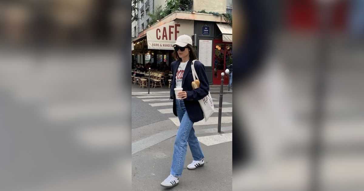 Mulher jovem, de bon&eacute; branco, &oacute;culos escuros, blazer azul-marinho, camiseta branca, jeans e t&ecirc;nis, atravessando a rua com uma sacola de p&atilde;o e um copo de caf&eacute; em frente a um caf&eacute; parisiense