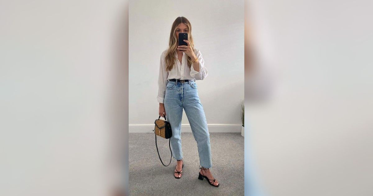 Mulher loira de p&eacute;, tirando selfie no espelho, vestindo camisa branca, cal&ccedil;a jeans clara de cintura alta, cinto preto, sand&aacute;lias pretas de salto baixo e bolsa de palha com al&ccedil;a preta