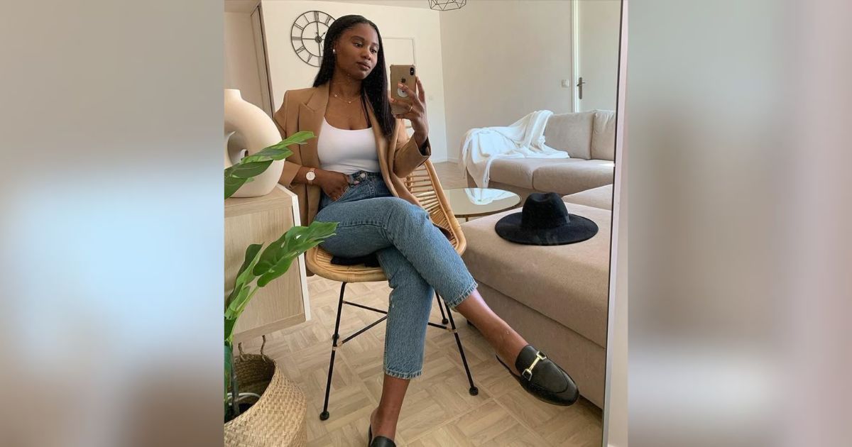 Mulher de pele escura, cabelo liso e comprido, sentada em uma cadeira de palha, tirando uma selfie. Ela veste blazer bege, top branco, jeans e mocassins pretos. Ao fundo, um espelho reflete um sof&aacute; bege e um chap&eacute;u preto. Uma planta e um vaso decoram o ambiente