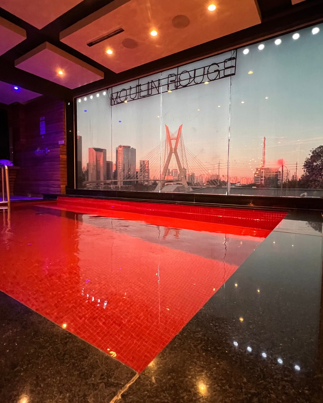 Piscina interna com azulejos vermelhos e borda preta, refletindo luzes do teto. Ao fundo, uma janela exibe a Ponte Estaiada e pr&eacute;dios de S&atilde;o Paulo ao p&ocirc;r do sol, com a palavra 