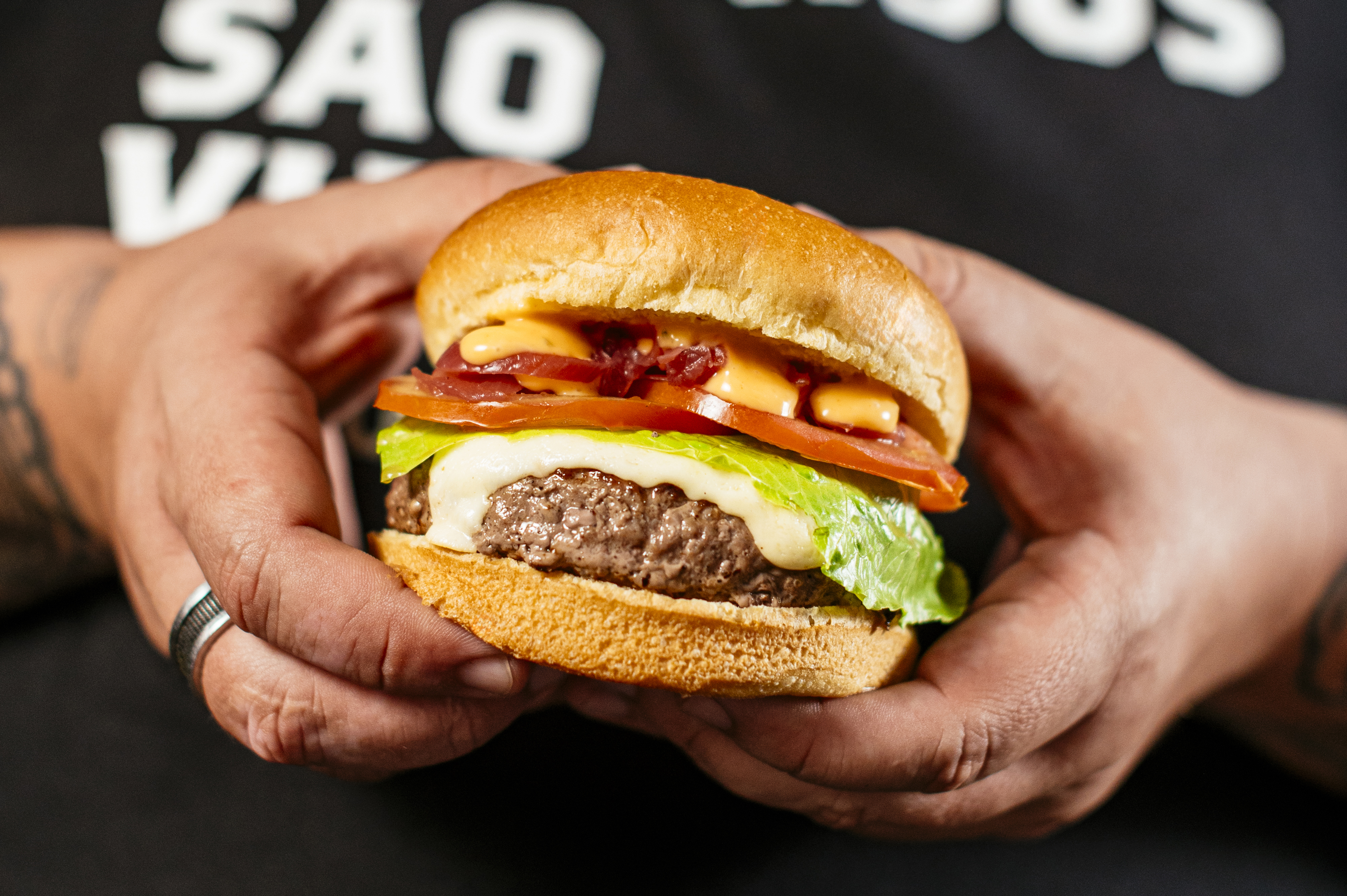 Mãos segurando um hambúrguer com pão dourado, carne, queijo derretido, alface, tomate e molho. Ao fundo, uma camiseta preta com a palavra "SÃO" em branco