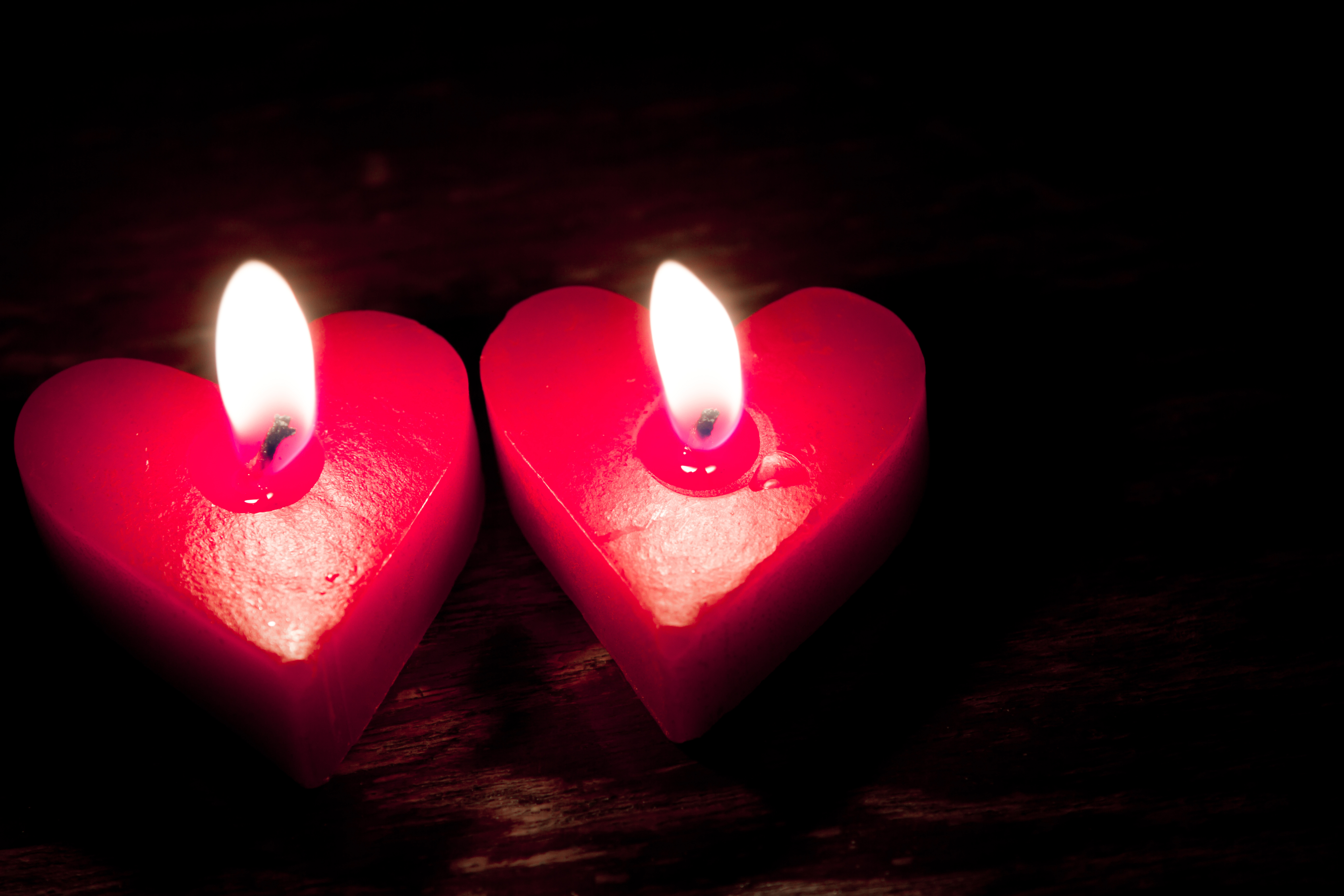 Duas velas vermelhas em formato de cora&ccedil;&atilde;o acesas, iluminando um fundo escuro de madeira, com a chama brilhante no centro de cada uma