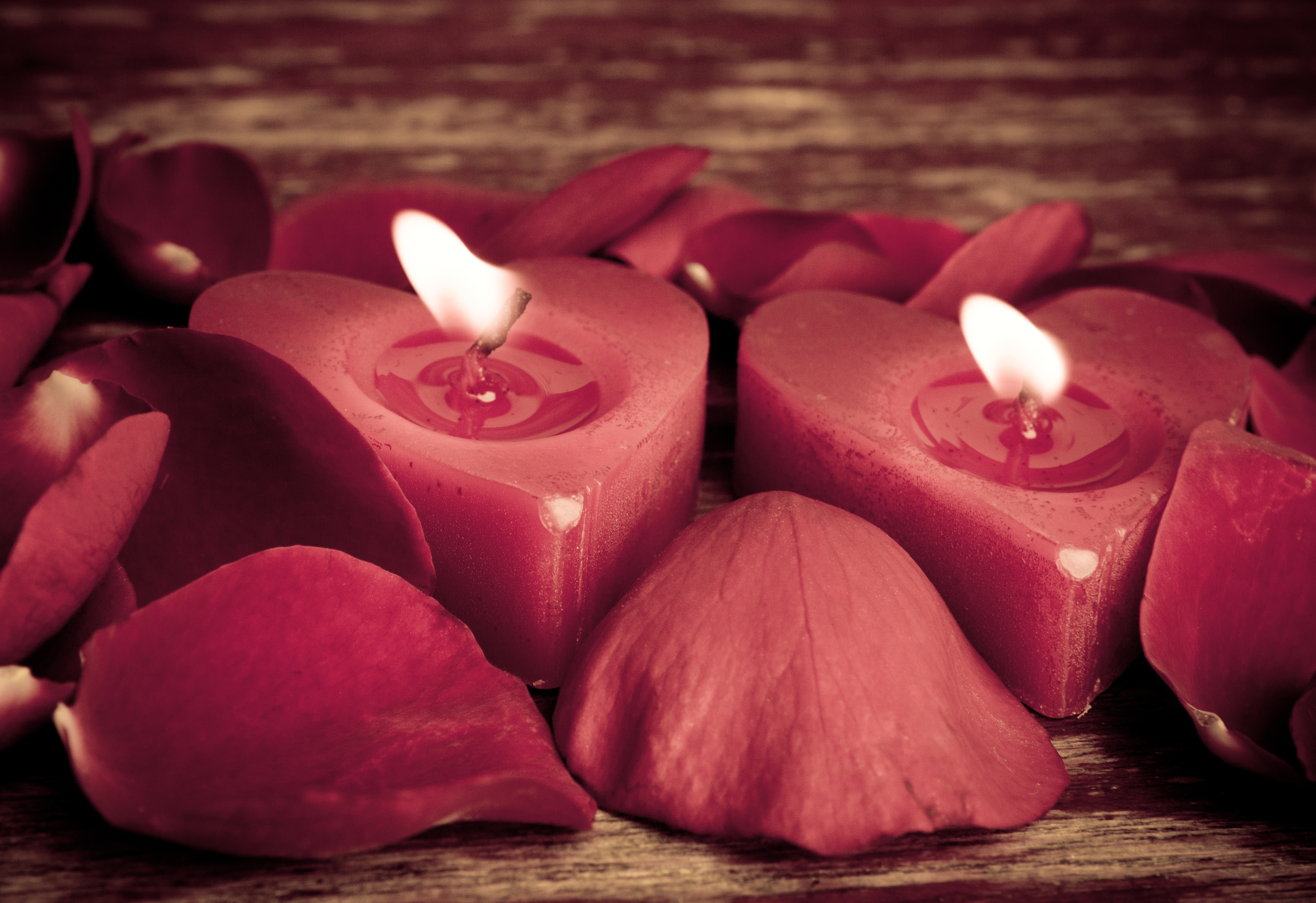Duas velas vermelhas em formato de cora&ccedil;&atilde;o acesas, cercadas por p&eacute;talas de rosa vermelhas, sobre uma superf&iacute;cie de madeira escura
