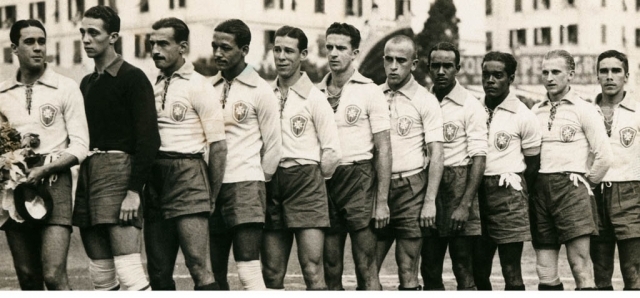 Onze jogadores de futebol, com idades e etnias variadas, posam em campo. O primeiro &agrave; esquerda segura um buqu&ecirc; de flores e um chap&eacute;u. Todos vestem camisas claras com distintivo no peito e shorts escuros, exceto um jogador de camisa escura. O fundo mostra um pr&eacute;dio e uma cerca com letras