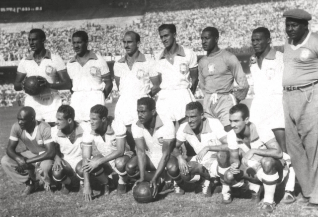 Time de futebol brasileiro posando para foto em campo, com 11 jogadores e um t&eacute;cnico. Seis jogadores est&atilde;o de p&eacute;, cinco agachados, e o t&eacute;cnico est&aacute; &agrave; direita, de p&eacute;. Todos vestem uniformes com escudo da sele&ccedil;&atilde;o, e alguns seguram bolas. A arquibancada lotada aparece ao fundo