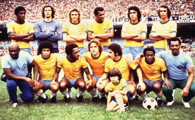 Time de futebol brasileiro posando para foto em campo de grama, com torcida ao fundo. Seis jogadores em p&eacute;, cinco agachados e uma crian&ccedil;a sentada, todos com camisas amarelas e azuis