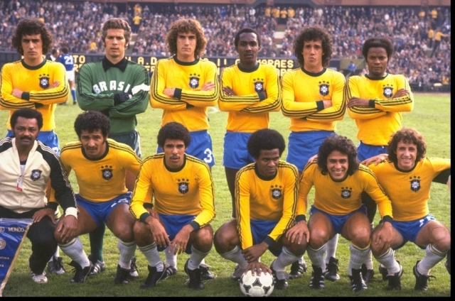 Time de futebol brasileiro posa para foto em campo, com 11 jogadores e um t&eacute;cnico. Seis jogadores est&atilde;o em p&eacute;, com camisas amarelas e cal&ccedil;&otilde;es azuis, bra&ccedil;os cruzados. Cinco jogadores est&atilde;o agachados na frente, com camisas amarelas e cal&ccedil;&otilde;es azuis, sorrindo. O t&eacute;cnico, &agrave; esquerda, veste jaqueta branca e preta. Ao fundo, arquibancada lotada