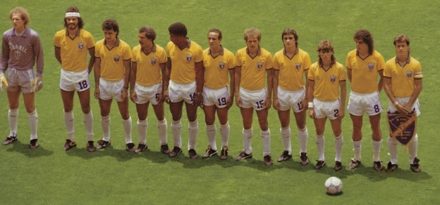 Time de futebol masculino com 11 jogadores em campo, um goleiro de roxo e os outros dez de camisa amarela e cal&ccedil;&atilde;o branco, posando para foto com uma bola no ch&atilde;o &agrave; direita