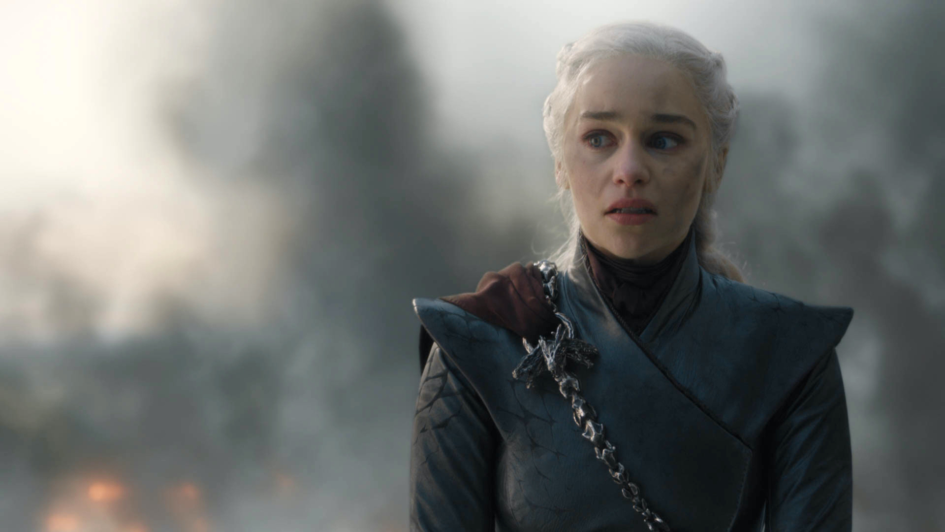 Daenerys Targaryen, com cabelos loiros platinados e tran&ccedil;ados, veste uma armadura escura com detalhes de drag&atilde;o no ombro. Seu rosto est&aacute; sujo, com express&atilde;o de choque e tristeza, olhando para o lado direito. O fundo est&aacute; emba&ccedil;ado, com fuma&ccedil;a e fogo, indicando um cen&aacute;rio de destrui&ccedil;&atilde;o