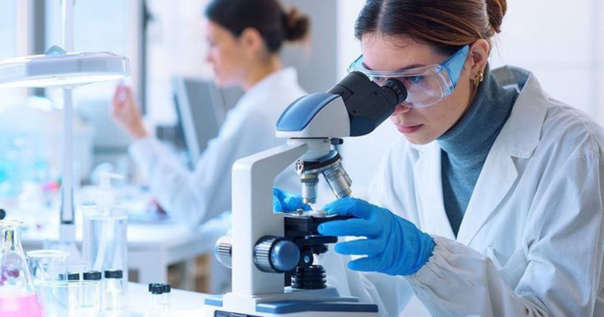 Cientista mulher de jaleco branco e luvas azuis, usando &oacute;culos de prote&ccedil;&atilde;o, observa amostra em microsc&oacute;pio em laborat&oacute;rio. Outra cientista trabalha ao fundo.