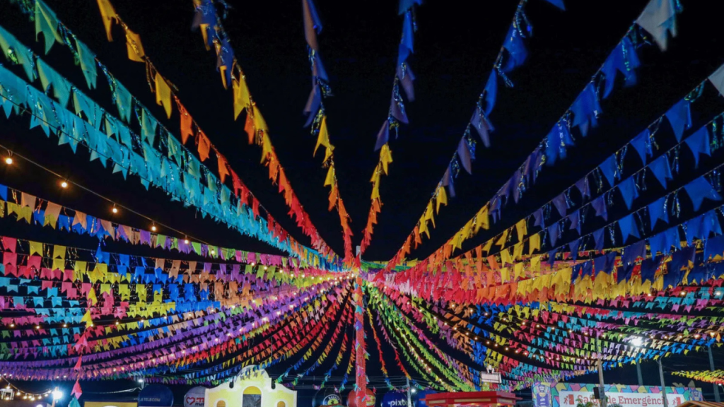 Céu noturno escuro com centenas de bandeirinhas coloridas de festa junina, em tons de azul, amarelo, vermelho, rosa e verde, penduradas em cordões que convergem para o centro. Pequenas lâmpadas incandescentes brilham entre as bandeirinhas, iluminando o ambiente festivo. Ao fundo, estruturas de barracas e decorações de festa junina são visíveis, com uma placa branca e vermelha indicando