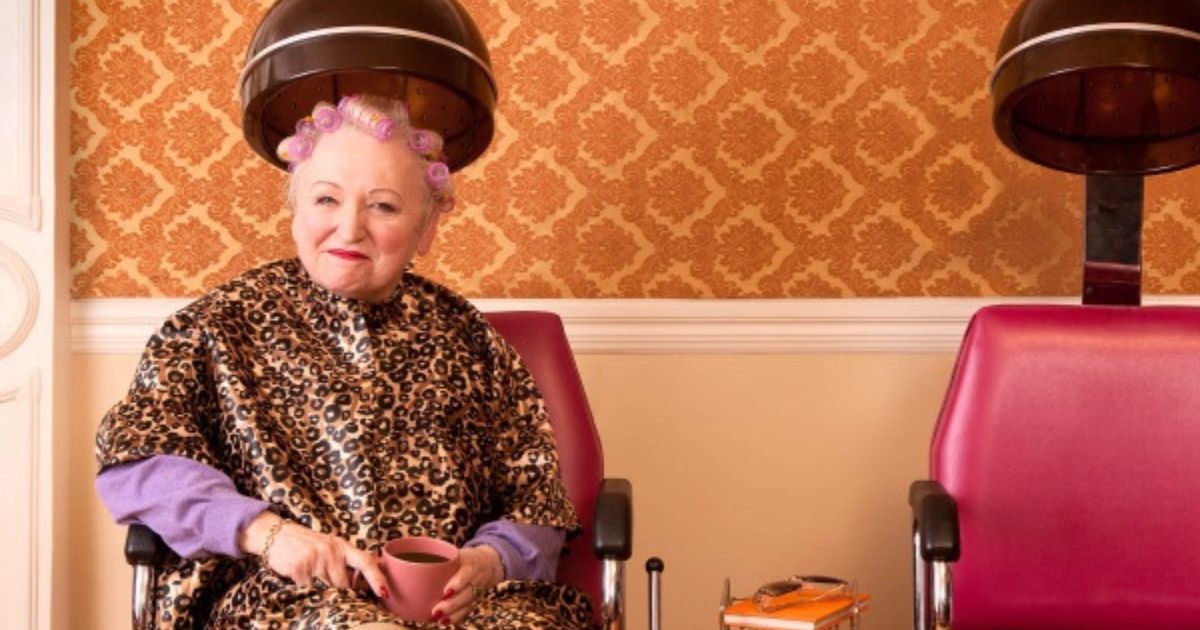 Mulher idosa sorridente, com bobes roxos no cabelo e capa de oncinha, sentada sob um secador de cabelo de sal&atilde;o, segurando uma x&iacute;cara rosa. Ao lado, uma cadeira de sal&atilde;o vazia e parede com papel de parede laranja estampado