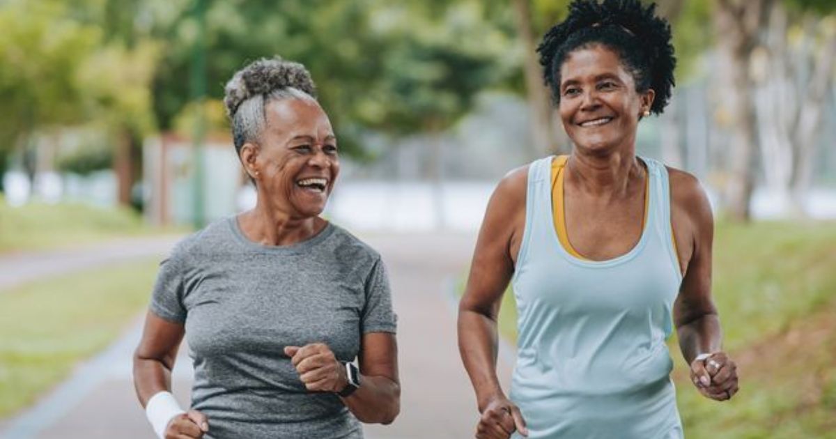 Duas mulheres idosas, sorrindo e correndo em um parque. A da esquerda veste camiseta cinza e rel&oacute;gio inteligente, a da direita, regata azul sobre top amarelo