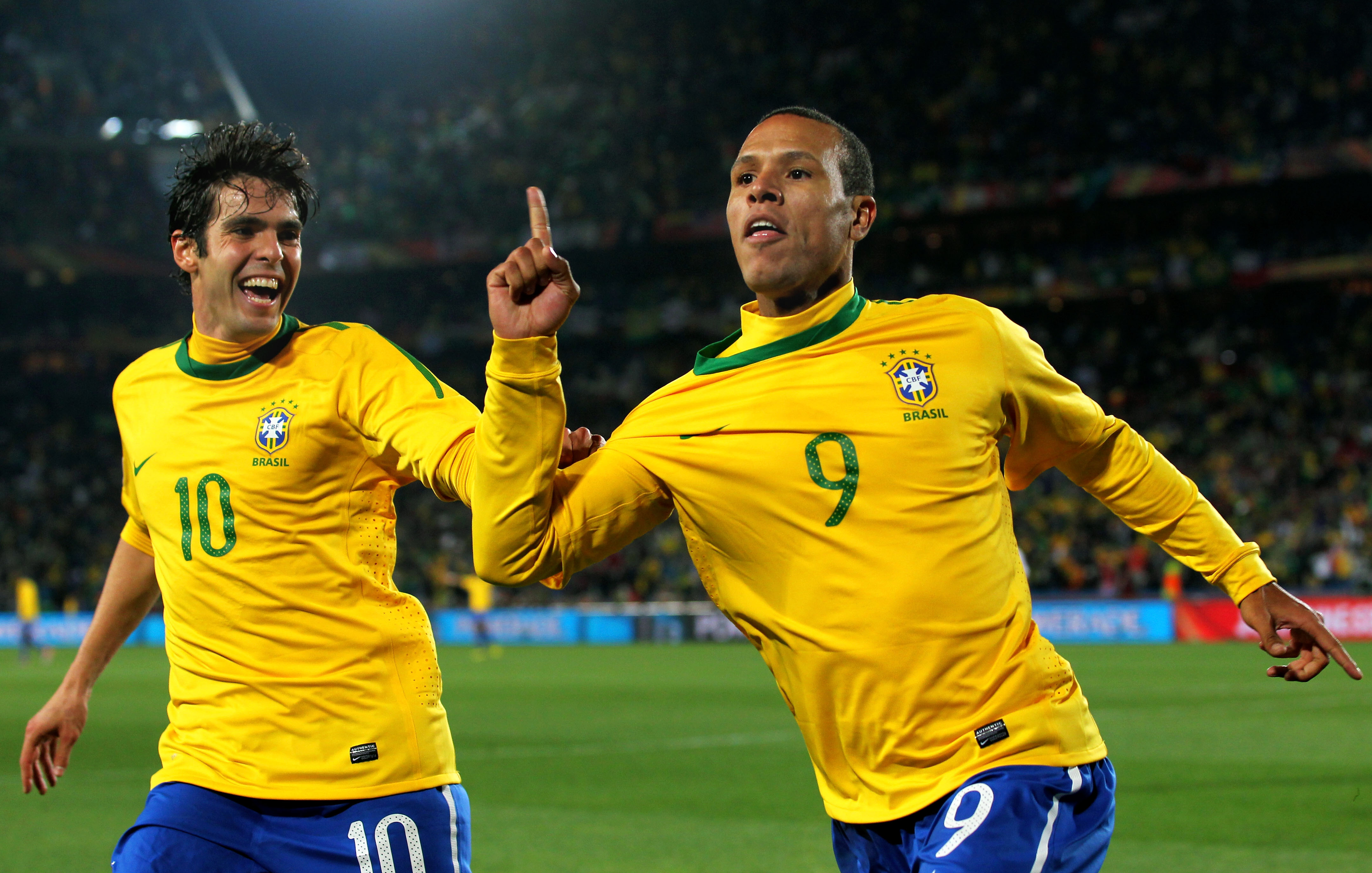 Dois jogadores de futebol brasileiros, Kak&aacute; e Lu&iacute;s Fabiano, comemoram um gol em campo. Kak&aacute;, &agrave; esquerda, sorri com a boca aberta, vestindo a camisa 10 amarela e azul. Lu&iacute;s Fabiano, &agrave; direita, com a camisa 9, aponta o dedo indicador para cima, ambos comemorando em um est&aacute;dio iluminado