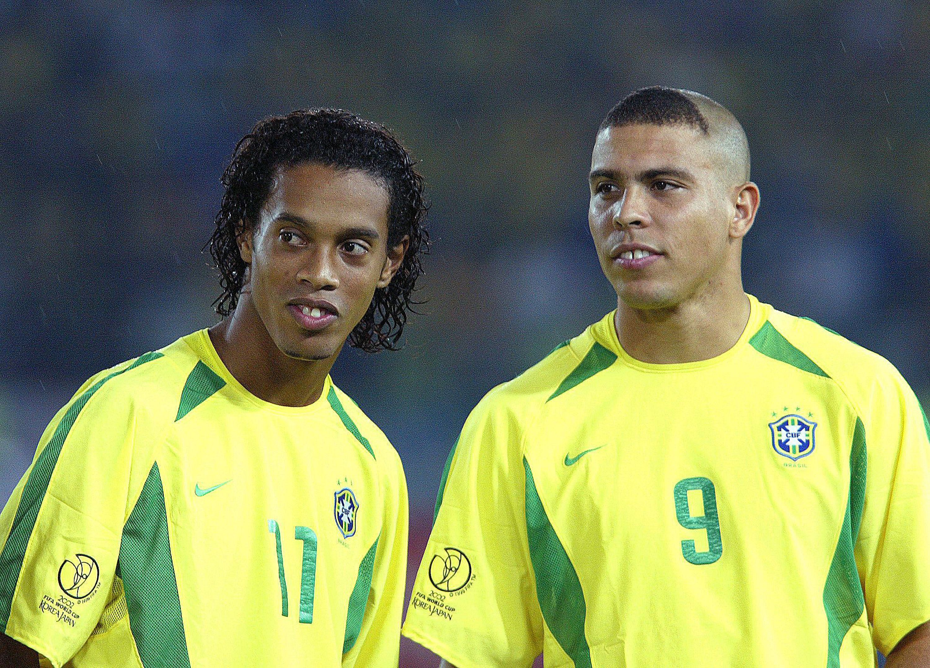 Ronaldinho Ga&uacute;cho, com cabelo encaracolado, e Ronaldo Fen&ocirc;meno, com corte de cabelo casc&atilde;o, vestem camisas amarelas da sele&ccedil;&atilde;o brasileira com detalhes verdes e o escudo da CBF. Ambos olham para a direita, com express&otilde;es s&eacute;rias, em um campo de futebol &agrave; noite