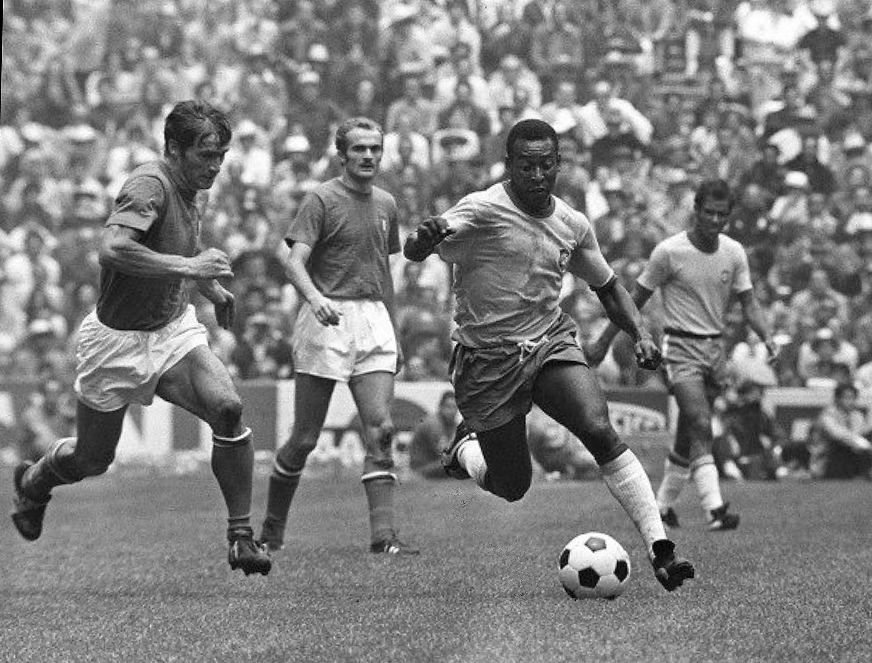 Pel&eacute;, de camisa clara e cal&ccedil;&atilde;o escuro, dribla a bola com o p&eacute; direito em campo de futebol, enquanto dois jogadores advers&aacute;rios, de camisas escuras e cal&ccedil;&otilde;es claros, o perseguem. A arquibancada lotada aparece ao fundo