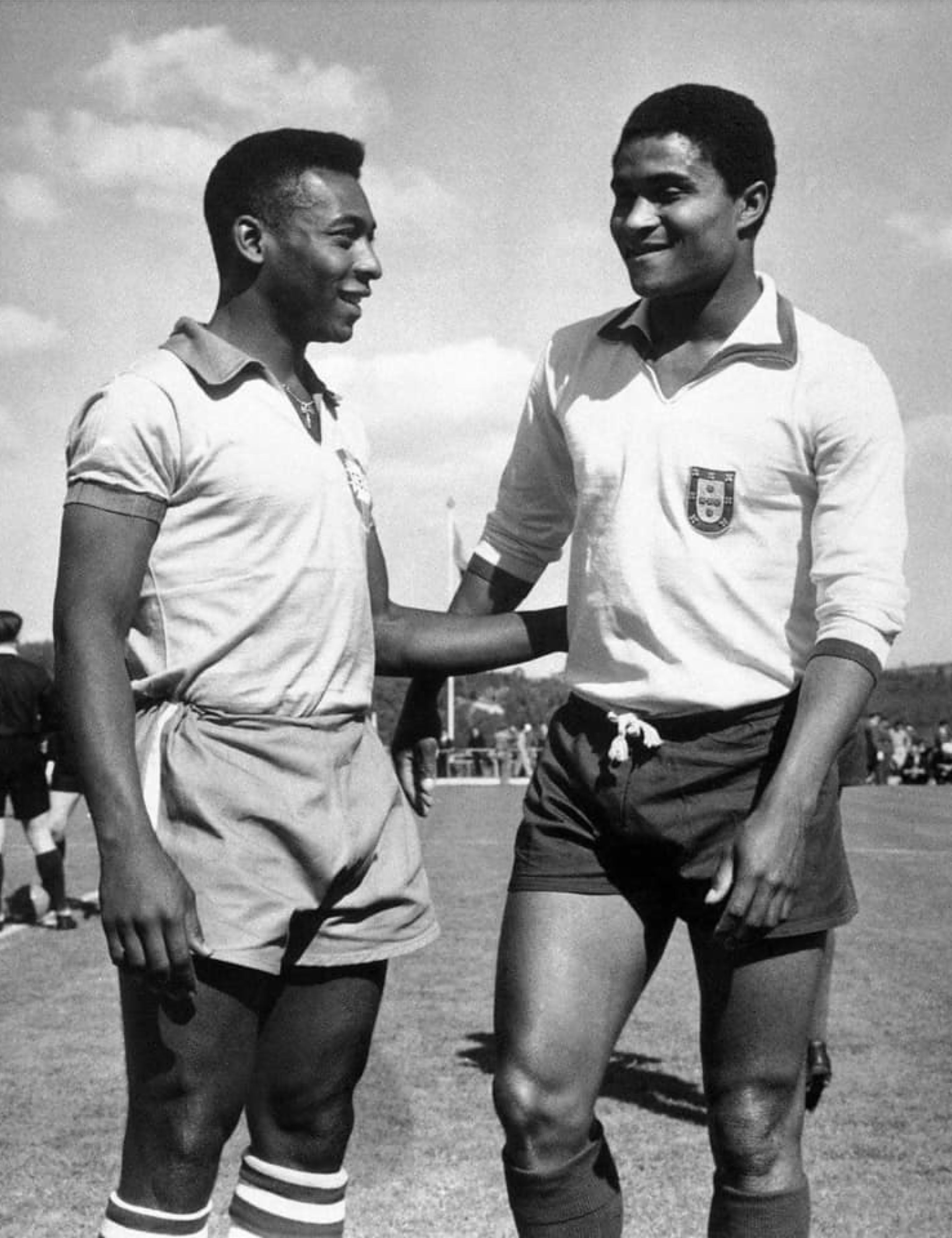 Pel&eacute; e Eus&eacute;bio, jogadores de futebol negros, sorriem um para o outro em campo. Pel&eacute; veste camisa clara de manga curta e shorts, Eus&eacute;bio camisa de manga comprida com escudo e shorts escuros. Pel&eacute; tem a m&atilde;o no ombro de Eus&eacute;bio.