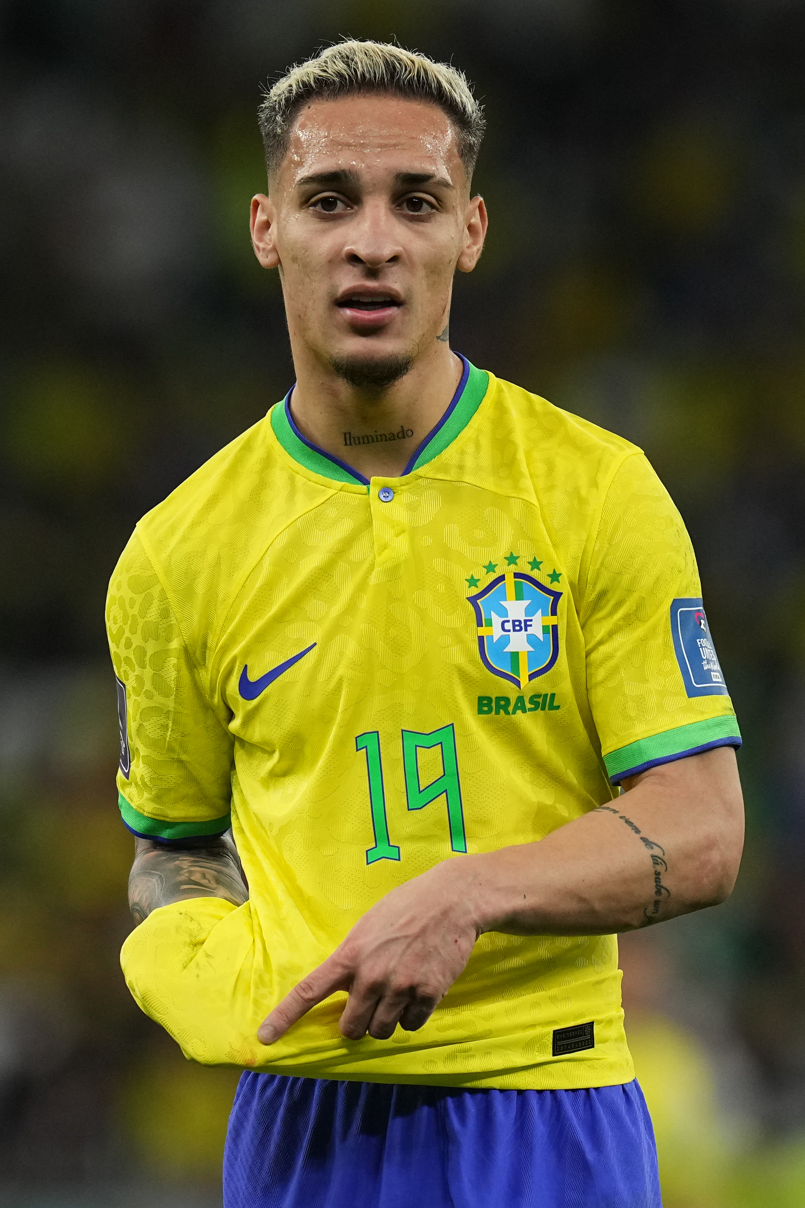 Jogador de futebol Antony, com cabelo loiro descolorido e tatuagens, vestindo a camisa amarela da sele&ccedil;&atilde;o brasileira com o n&uacute;mero 19 em verde e o distintivo da CBF. Ele aponta para baixo com a m&atilde;o direita, em um campo de futebol.