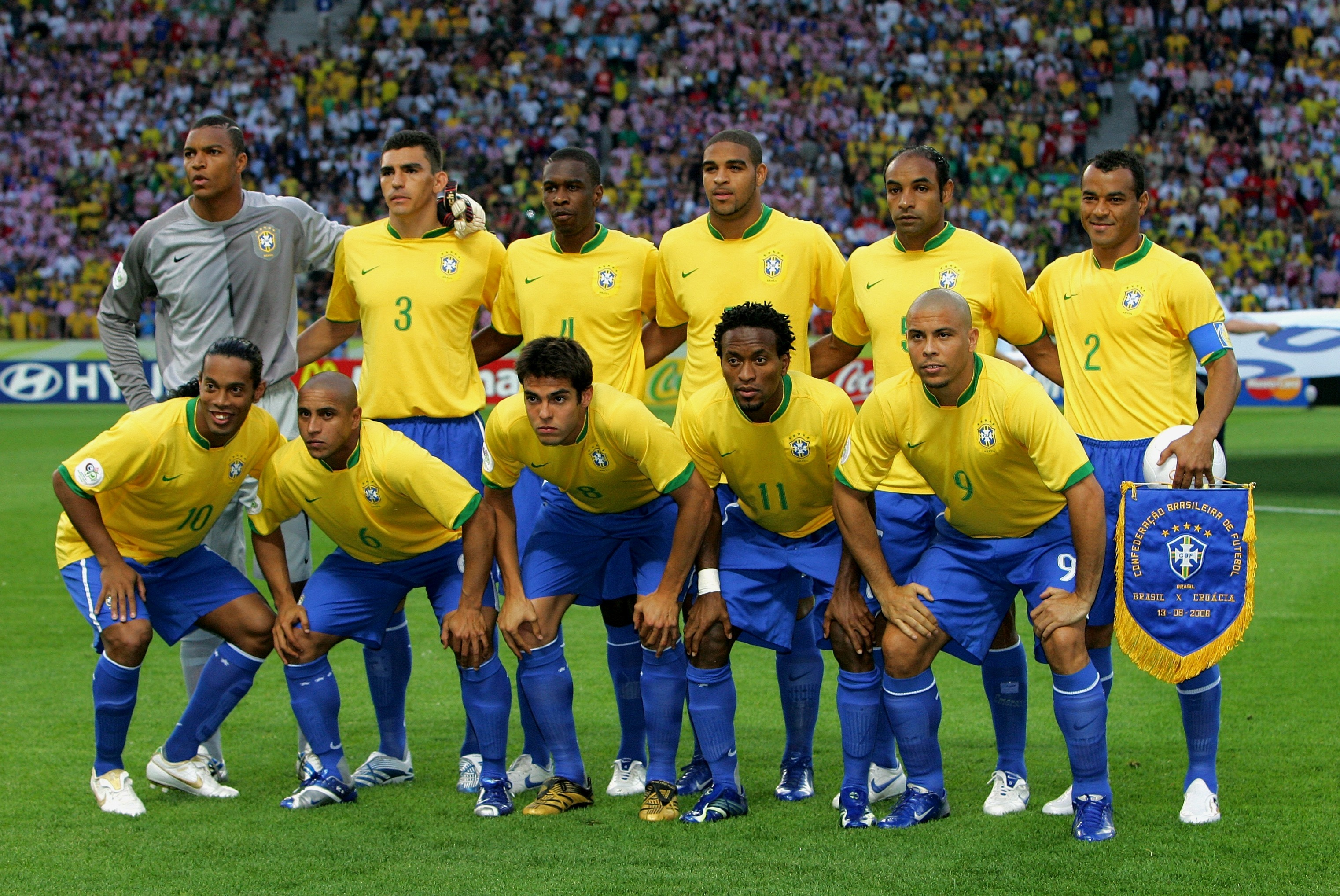 Jogadores da sele&ccedil;&atilde;o brasileira de futebol, com camisas amarelas e shorts azuis, posam para foto em campo de grama verde, com torcida ao fundo. Onze atletas, alguns em p&eacute; e outros agachados, exibem um estandarte azul com o escudo da Confedera&ccedil;&atilde;o Brasileira de Futebol
