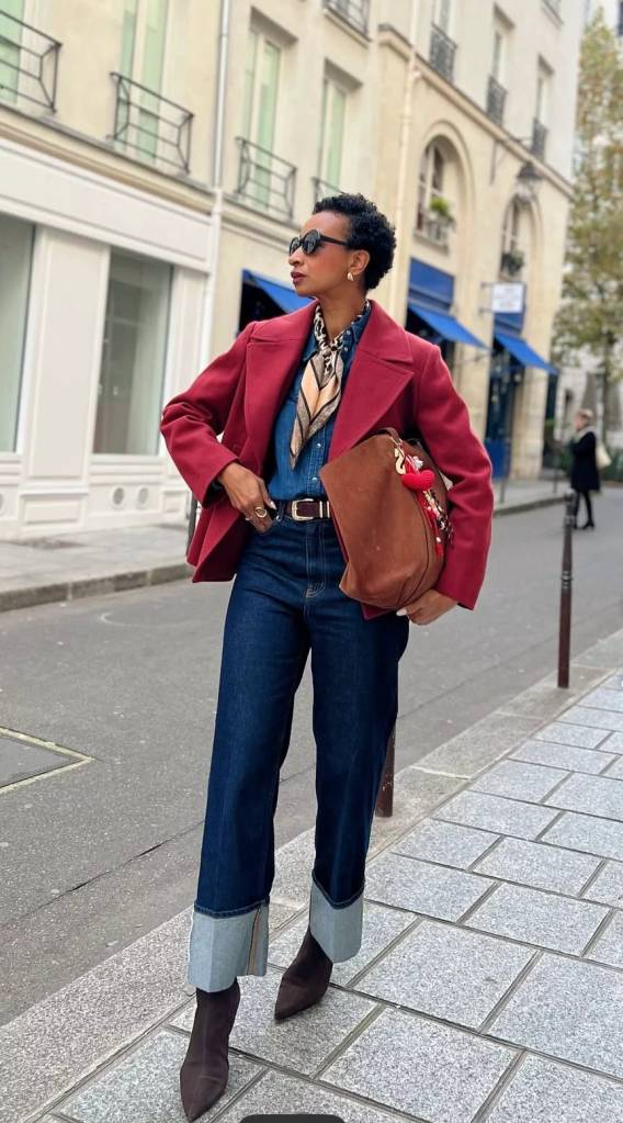 Mulher negra de cabelo curto e &oacute;culos escuros, usando blazer vinho, camisa jeans, cal&ccedil;a jeans escura com barra virada e botas marrons, segurando uma bolsa de camur&ccedil;a marrom, em uma rua de paralelep&iacute;pedos com pr&eacute;dios claros ao fundo