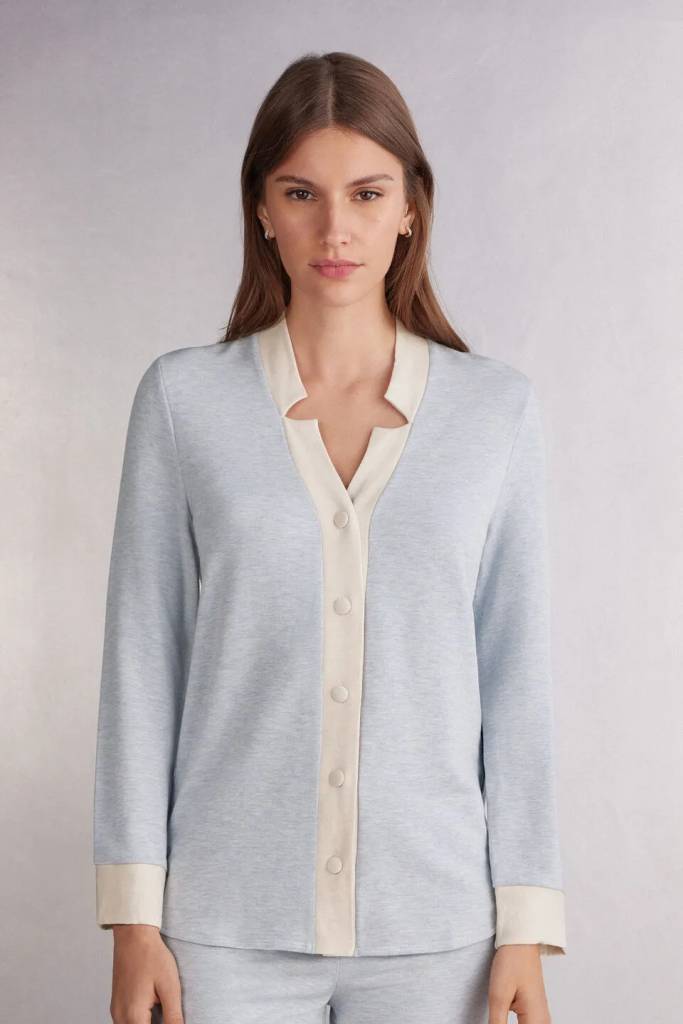 Mulher de cabelos castanhos longos, com express&atilde;o s&eacute;ria, vestindo um conjunto de pijama azul claro com detalhes em creme, incluindo gola, bot&otilde;es e punhos. Ela usa brincos pequenos e est&aacute; em um fundo cinza claro