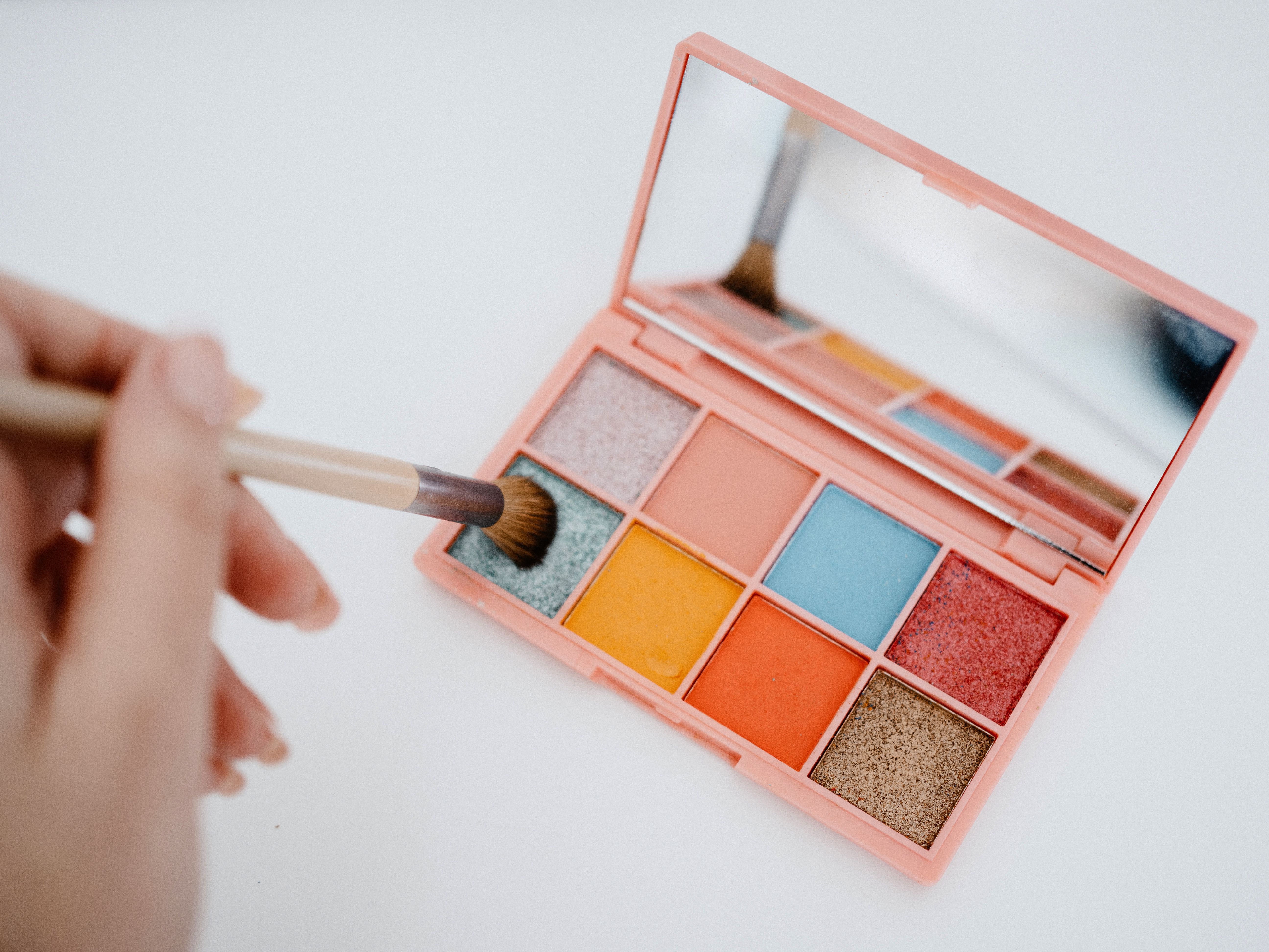 Mão feminina segurando um pincel de maquiagem sobre uma paleta de sombras rosa aberta, com espelho e oito cores vibrantes, incluindo azul, laranja, dourado e tons cintilantes