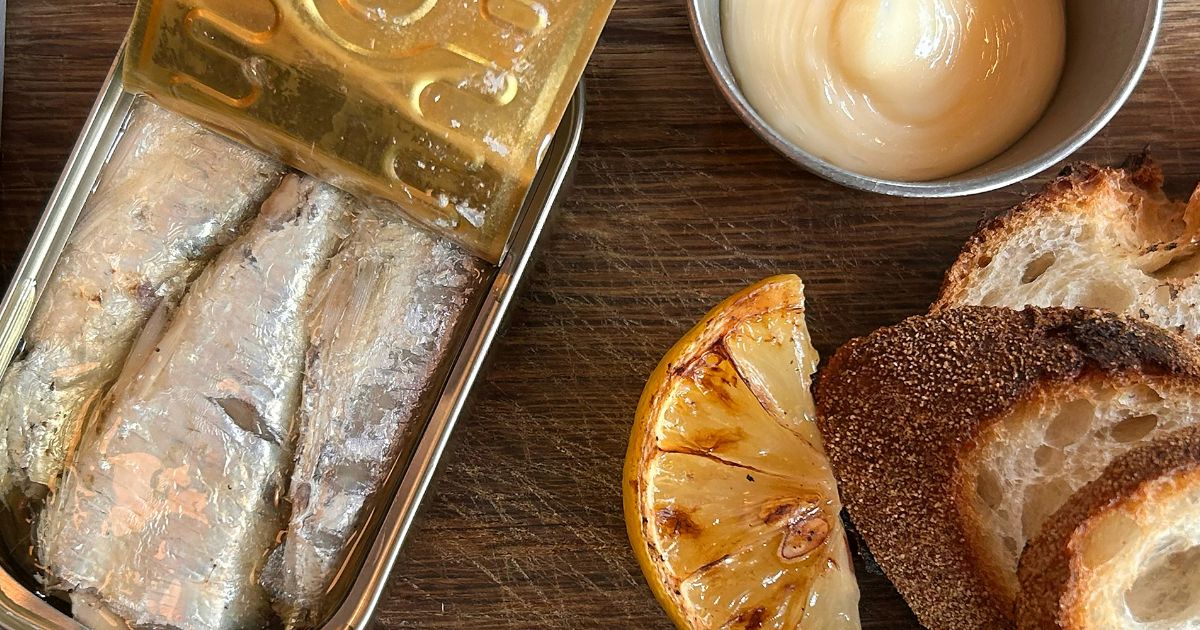 Lata de sardinhas abertas com peixes prateados, fatias de p&atilde;o r&uacute;stico, um gomo de lim&atilde;o grelhado e um pote de molho cremoso sobre uma t&aacute;bua de madeira
