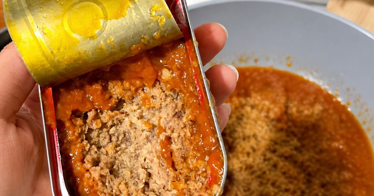 Uma m&atilde;o segura uma lata de sardinha aberta, revelando o peixe desfiado em molho vermelho. Ao fundo, um prato com mais molho e temperos.