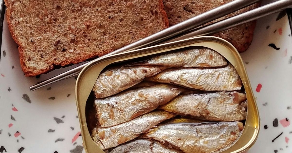 Uma lata aberta de sardinhas em conserva, com peixes prateados empilhados, ao lado de duas fatias de p&atilde;o integral com gr&atilde;os. Um par de hashis met&aacute;licos repousa sobre a lata e o p&atilde;o, em um prato branco com detalhes em preto e rosa