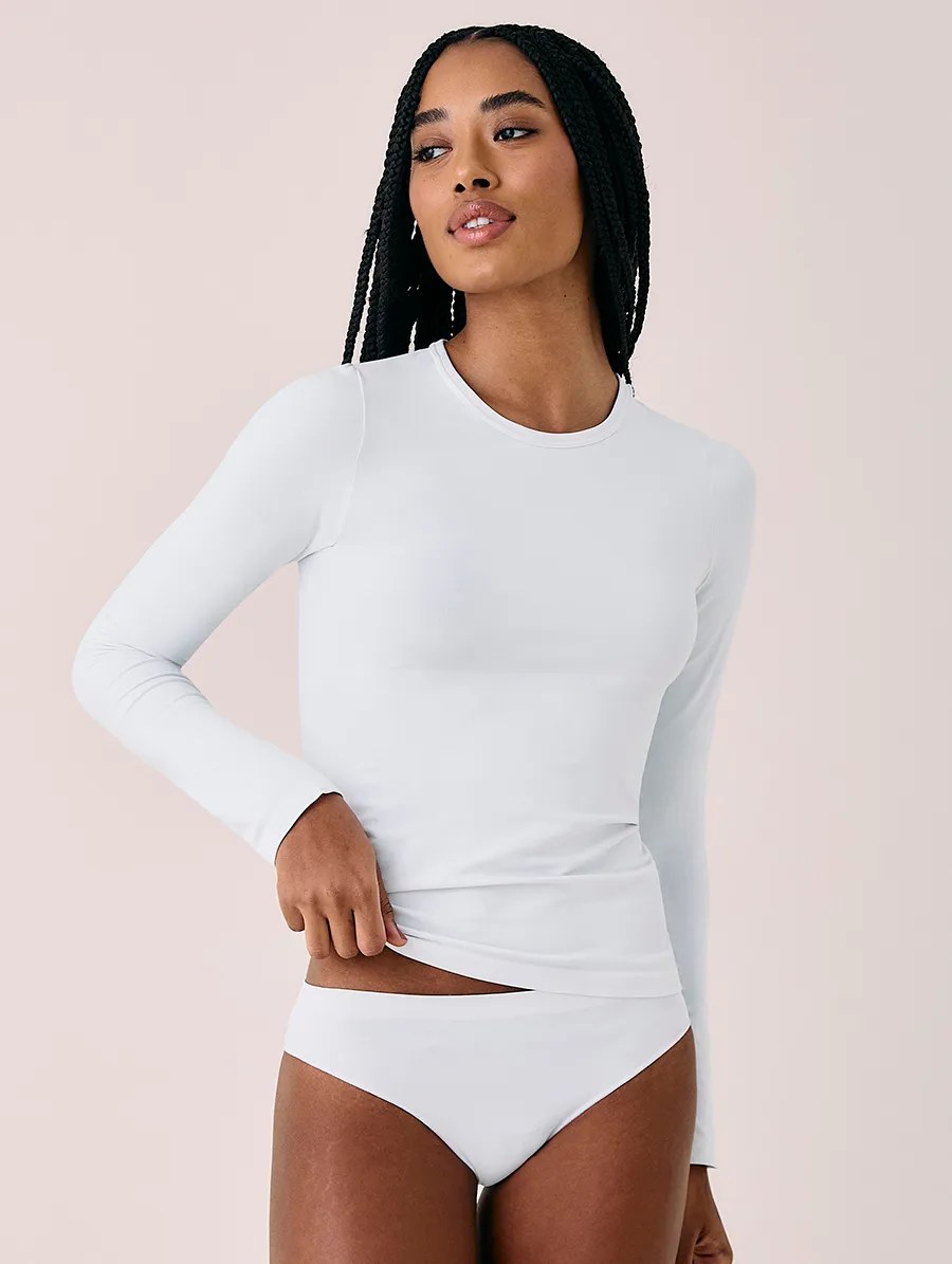 Mulher negra com tran&ccedil;as longas, vestindo camiseta branca de manga comprida e calcinha branca, puxa a barra da camiseta com a m&atilde;o direita, olhando para a direita com express&atilde;o serena, em fundo rosa claro