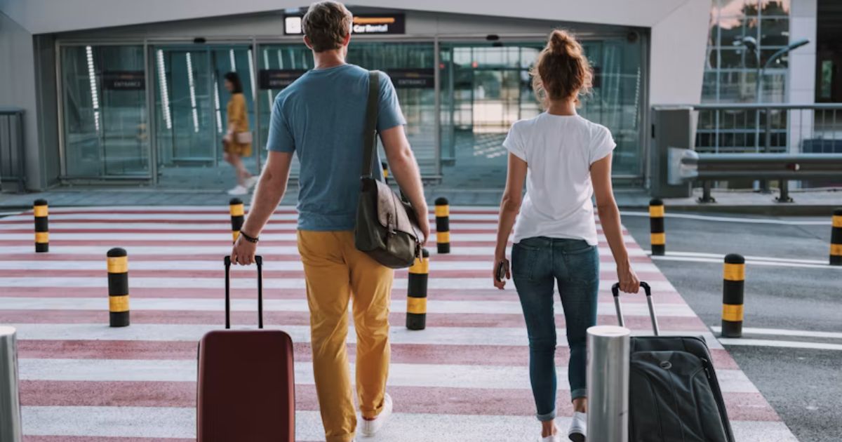 Casal, visto de costas, atravessando uma faixa de pedestres vermelha e branca com malas de rodinha, em direção a um prédio moderno com portas de vidro, possivelmente um aeroporto