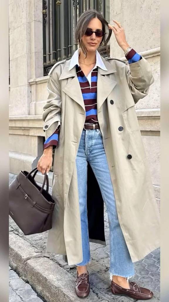 Mulher com &oacute;culos escuros, casaco bege, camisa listrada azul e vinho, jeans e sapatos marrons, segurando uma bolsa escura, em frente a um pr&eacute;dio de pedra