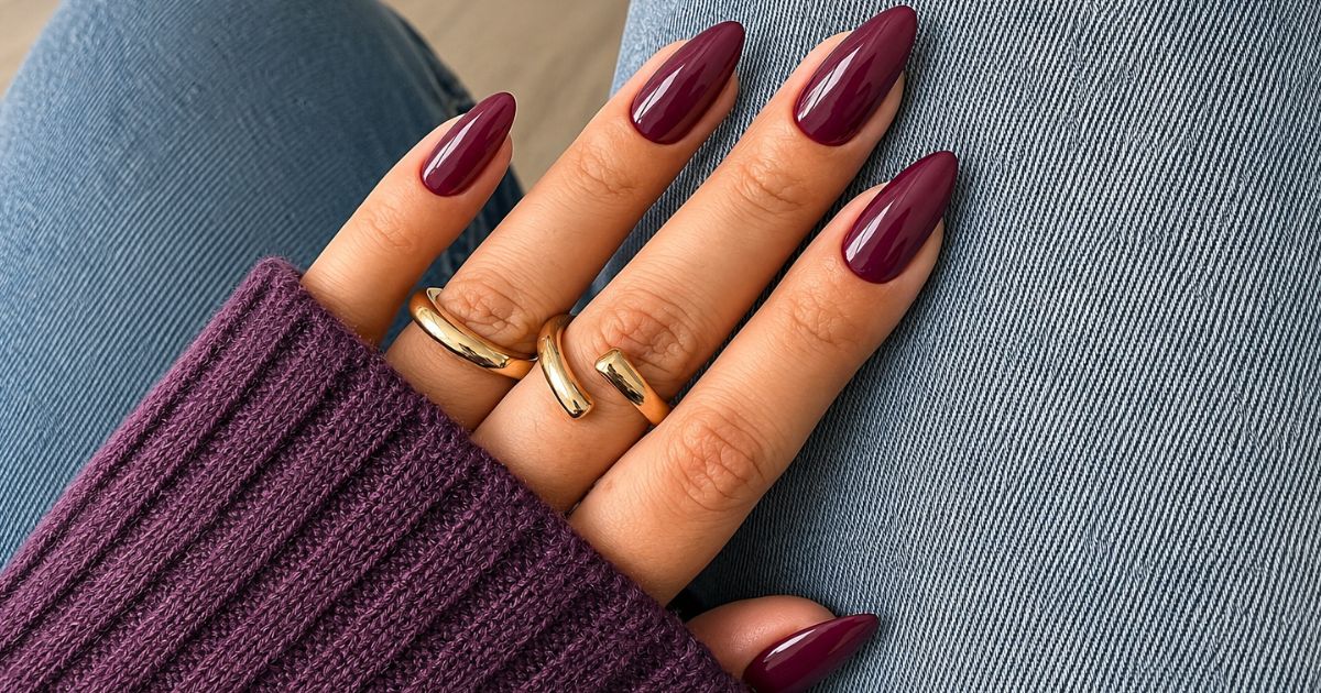 M&atilde;o feminina com unhas longas e pontiagudas pintadas de vinho, usando dois an&eacute;is dourados minimalistas, sobre uma manga de su&eacute;ter roxo e cal&ccedil;a jeans azul