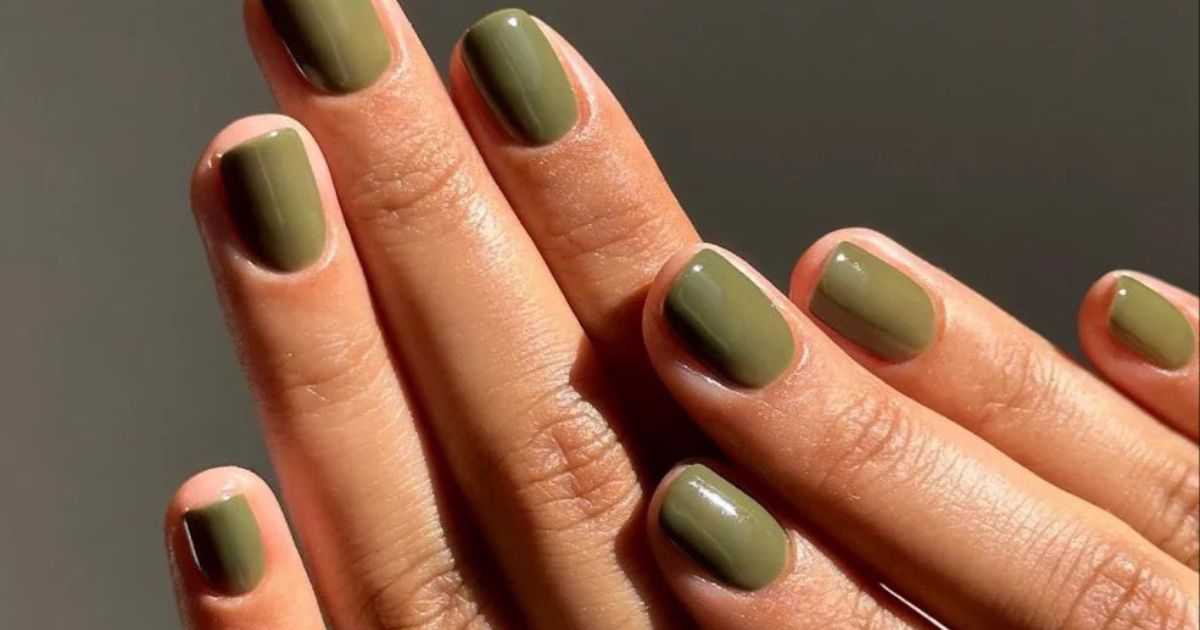 M&atilde;os com unhas curtas e arredondadas, pintadas com esmalte verde-oliva brilhante, em um fundo cinza escuro. A luz incide sobre os dedos, destacando o brilho do esmalte e a textura da pele