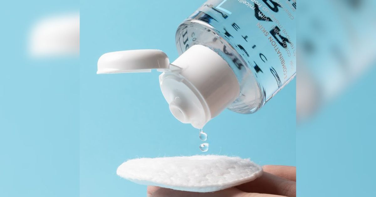 Uma garrafa transparente de &aacute;gua micelar com tampa branca aberta derramando duas gotas em um disco de algod&atilde;o branco, segurado por uma m&atilde;o, sobre um fundo azul claro