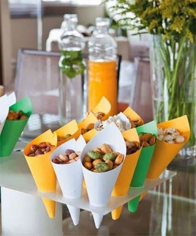 Cones de papel coloridos, brancos, amarelos e verdes, cheios de amendoins e castanhas variadas, dispostos em um suporte de acr&iacute;lico. Ao fundo, garrafas de suco de laranja e &aacute;gua com hortel&atilde;, e um vaso de flores amarelas, sugerindo um evento ou festa