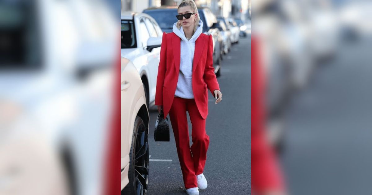 Mulher loira, com rabo de cavalo e &oacute;culos escuros, usando blazer e cal&ccedil;a vermelhos sobre um moletom branco, caminhando na rua com carros estacionados ao fundo