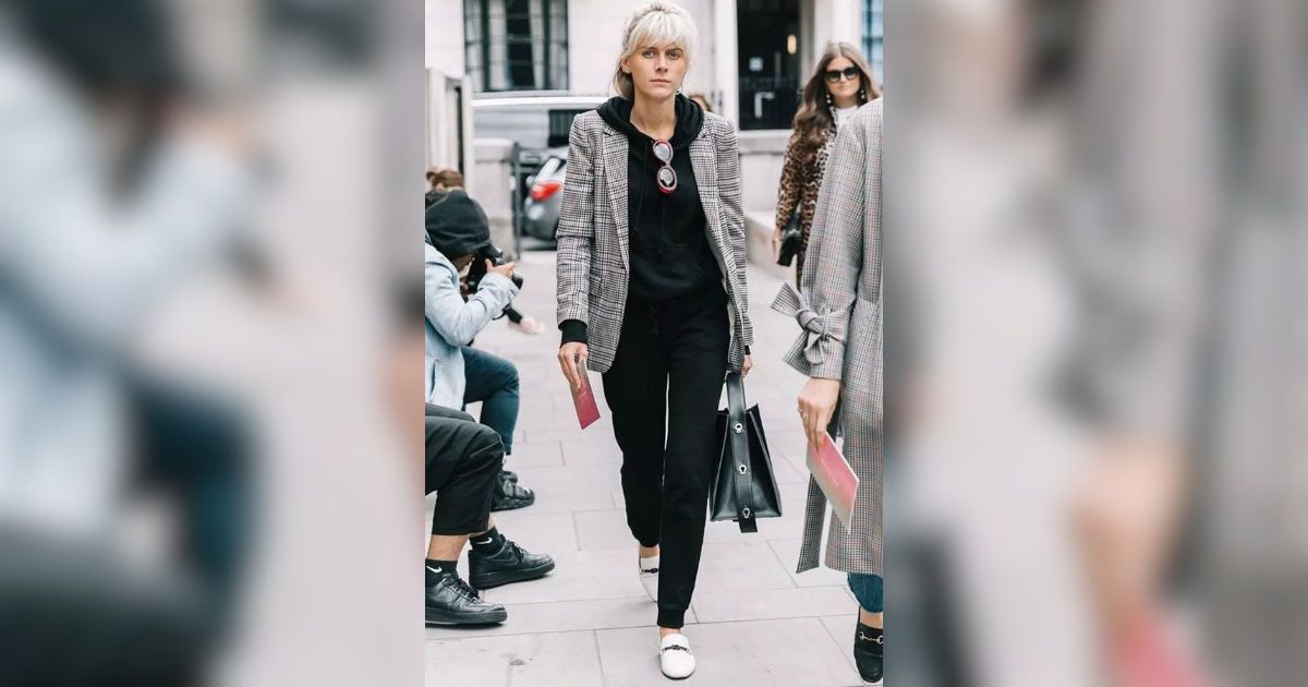 Mulher loira de cabelo preso, blazer xadrez cinza e preto sobre moletom preto, cal&ccedil;a preta e sapatos brancos, caminhando em cal&ccedil;ada urbana com bolsa preta na m&atilde;o esquerda e um papel rosa na direita. Outras pessoas desfocadas ao fundo.