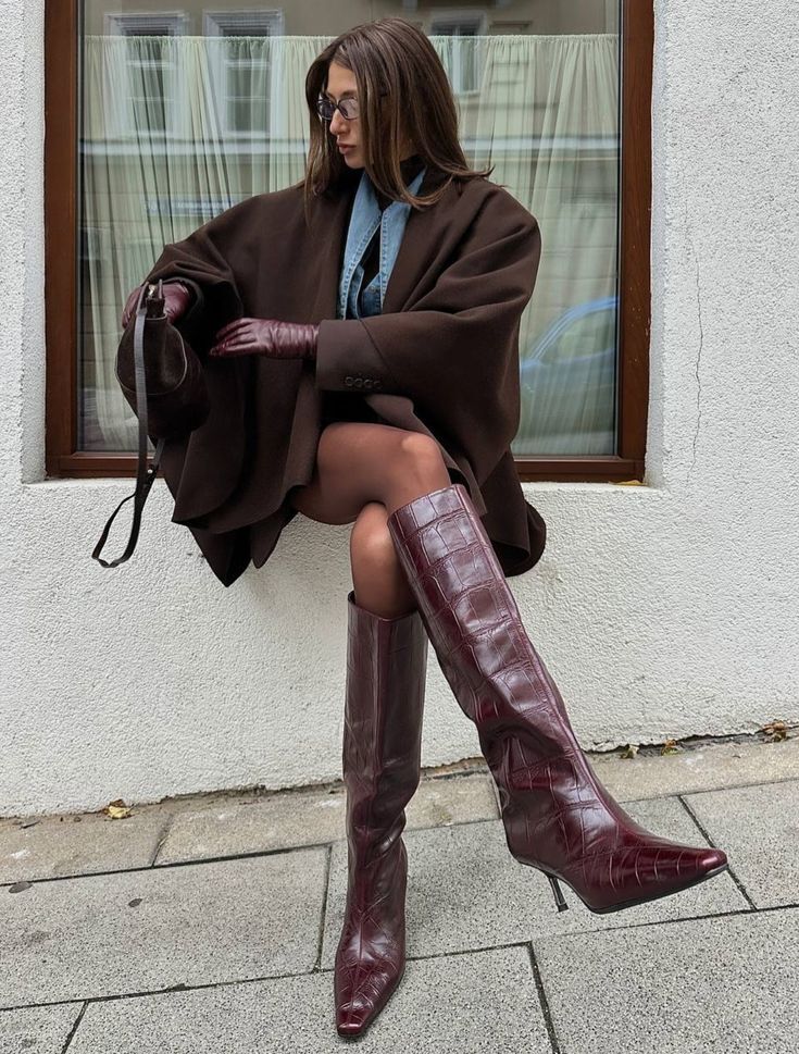 Mulher de &oacute;culos, cabelos castanhos e compridos, sentada em um parapeito branco, vestindo casaco marrom, camisa jeans, luvas e botas de cano alto cor vinho, segurando uma bolsa pequena marrom. O fundo mostra uma janela com cortina branca e uma cal&ccedil;ada cinza