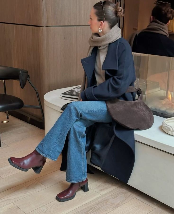 Mulher jovem sentada de perfil, usando casaco azul-marinho, su&eacute;ter cinza, cachecol bege, jeans e botas de couro vinho. Ela tem o cabelo preso e brincos prateados. Ao fundo, uma parede de madeira e uma lareira com fogo.