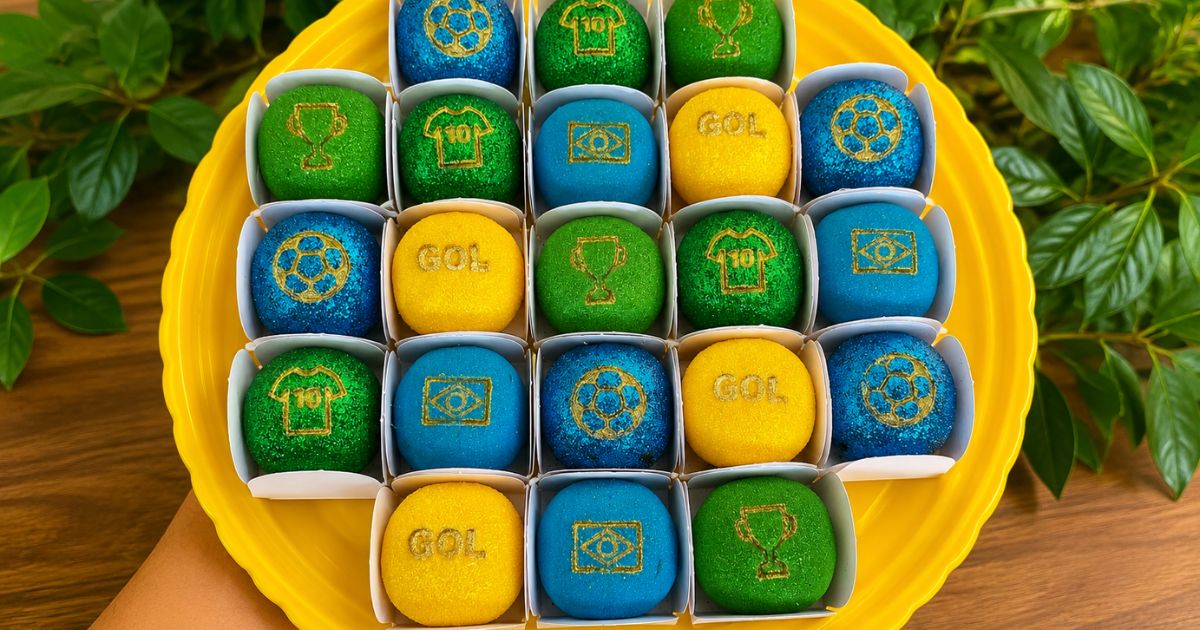Uma bandeja amarela com 25 docinhos redondos em forminhas brancas, nas cores verde, azul e amarelo, com símbolos dourados de futebol como troféu, camisa 10, bola e a palavra GOL